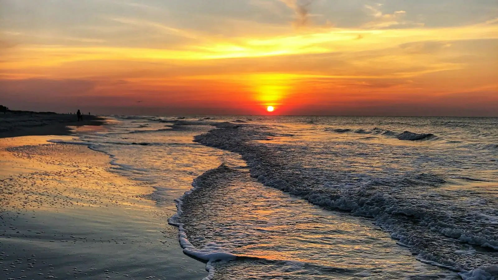 Low waves running up a wide, flat stretch of pale sand in late evening light