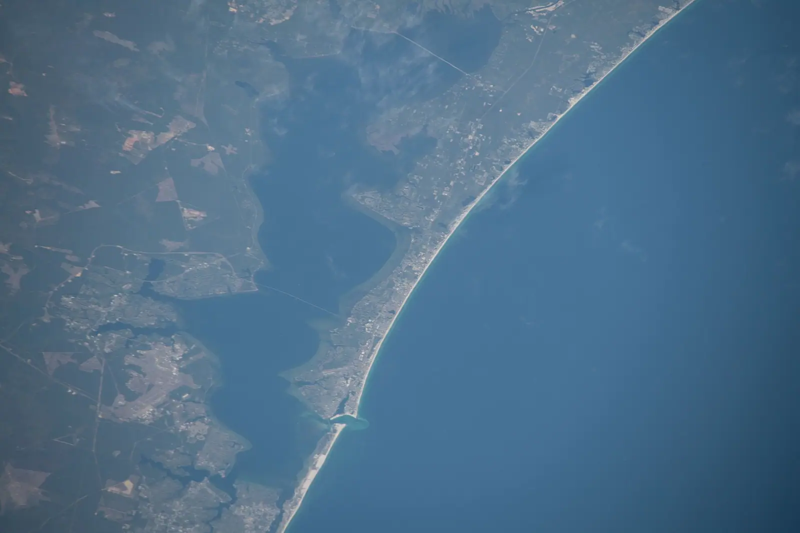 An aerial view of a long low bridge crossing shallow green water with a pale sandbar visible beneath the surface