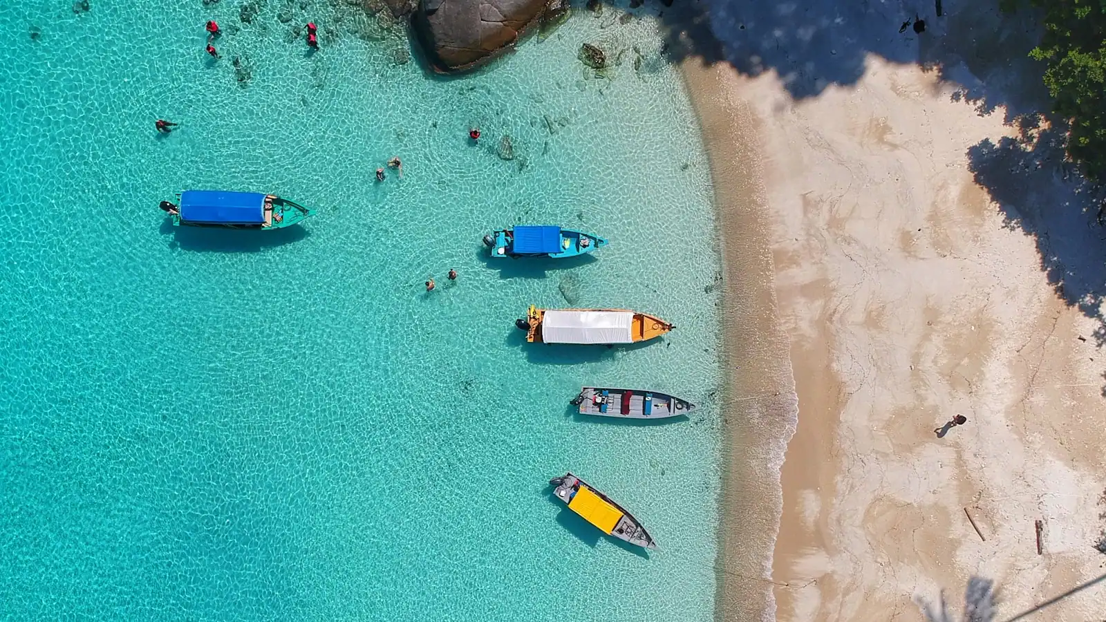 Boats moored together over clear shallow water beside a sandbank, seen from the air