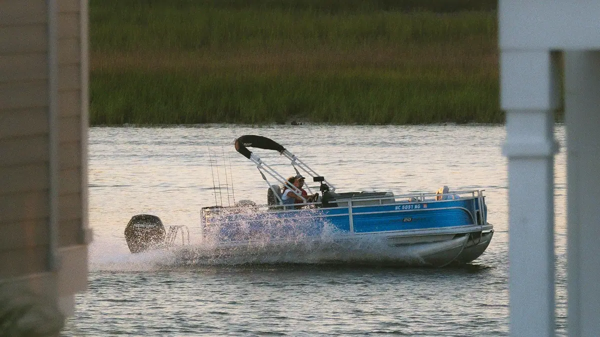 A blue pontoon boat under way on flat water past a low waterfront shoreline