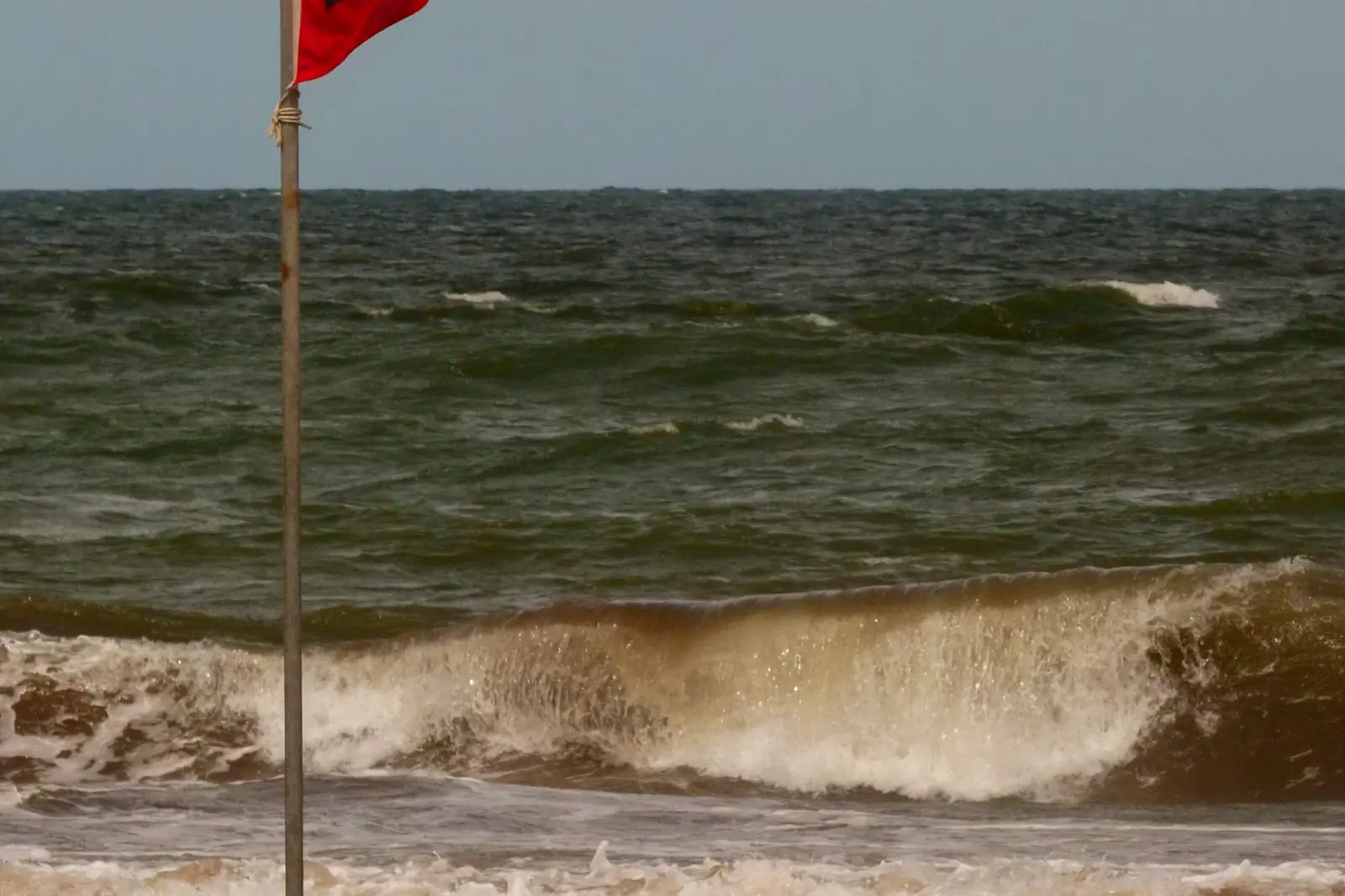A red warning flag flying from a pole at the edge of a beach with surf behind it