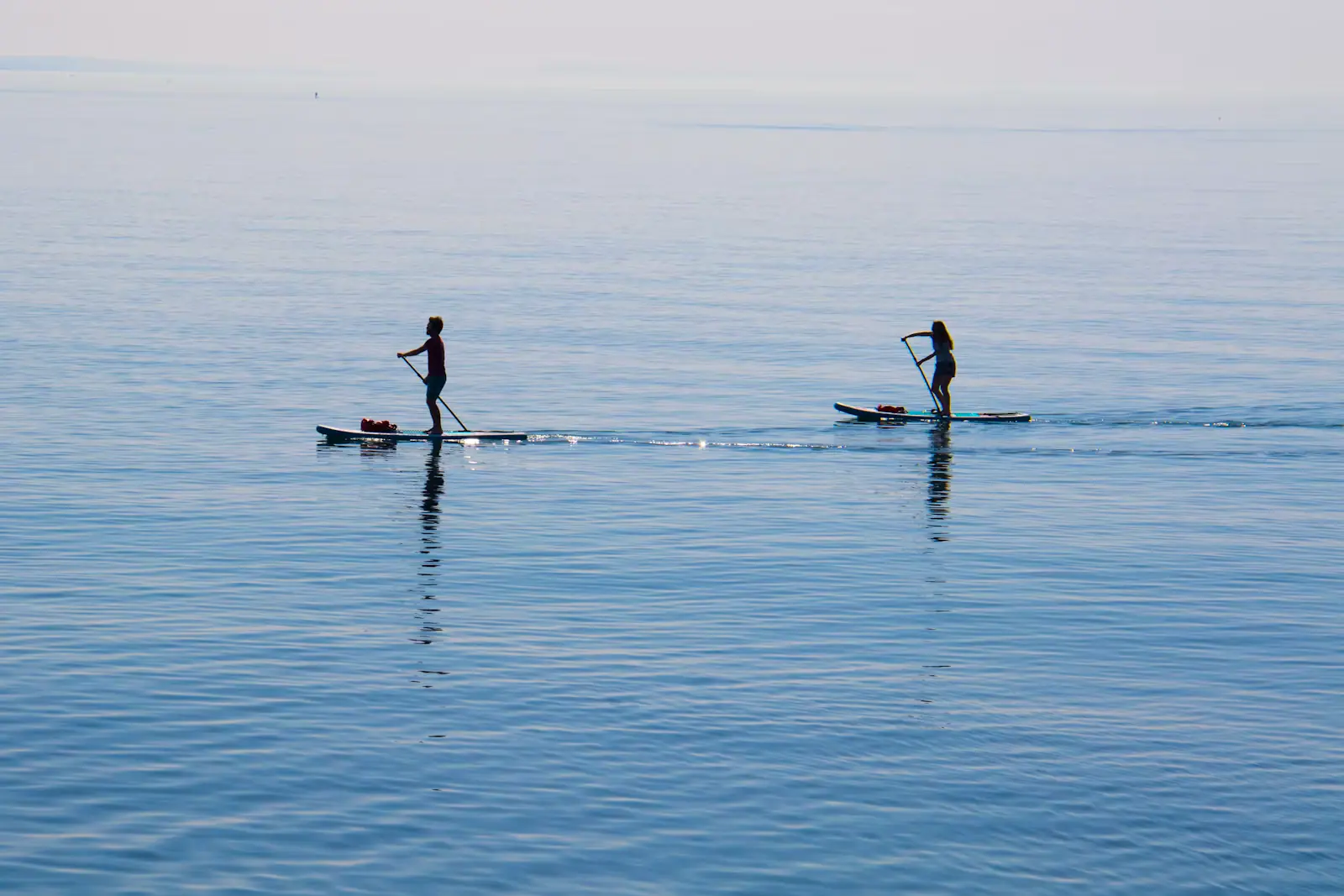Two paddleboarders on a wide sheet of flat, pale, mirror-calm water under a soft sky