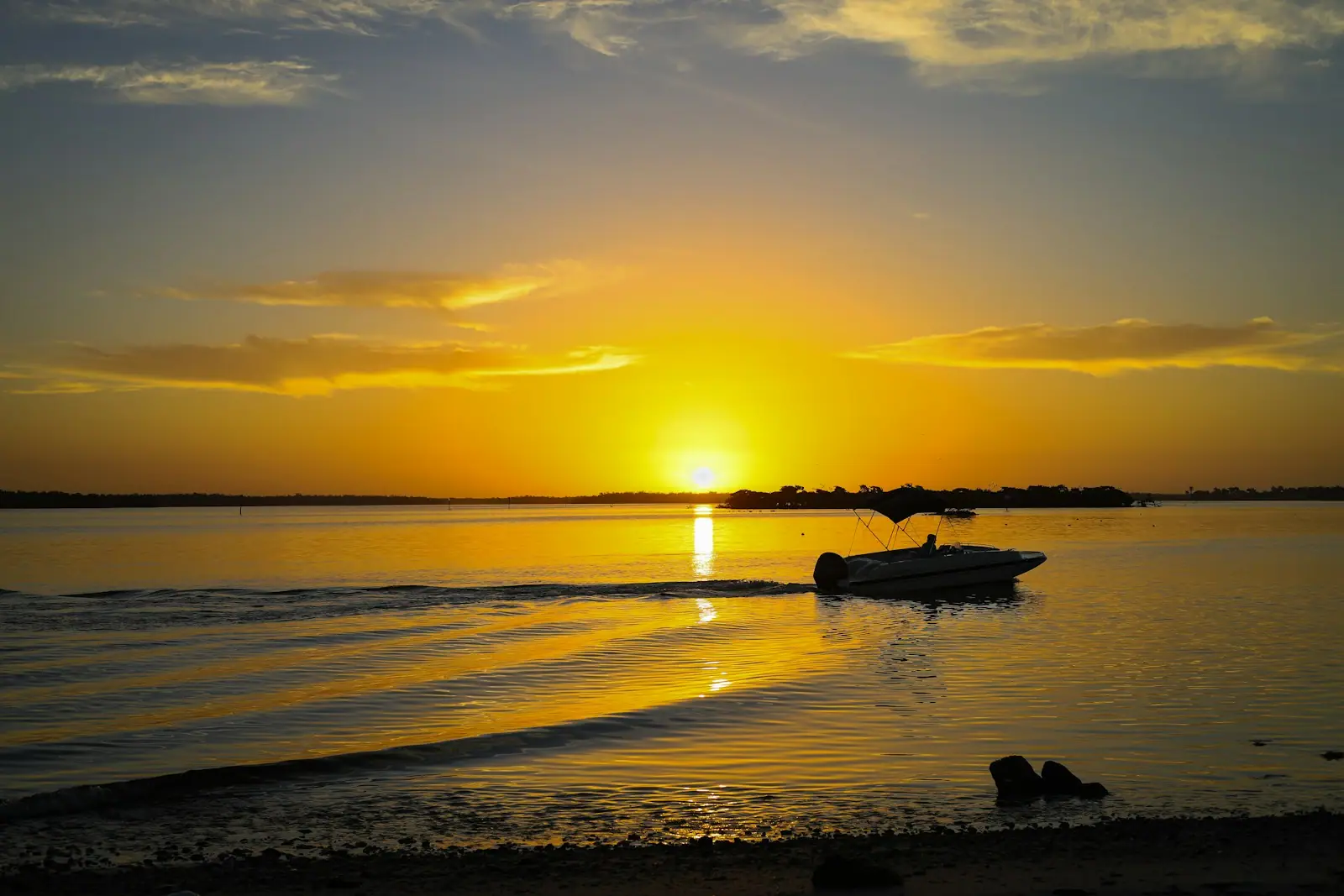 The sun setting over calm water with a small boat silhouetted near the shoreline